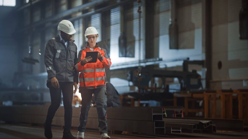 A male and female contractor standing side-by-side as they walk and discuss what they are viewing on a tablet.